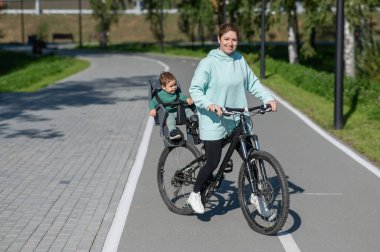 Küçük oğluyla bisiklete binen beyaz bir kadın arkasında çocuk koltuğunda oturuyor.
