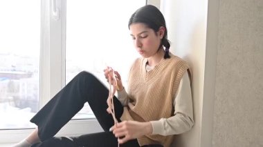 Genç beyaz kadın pencere eşiğinde otururken örgü örüyor.