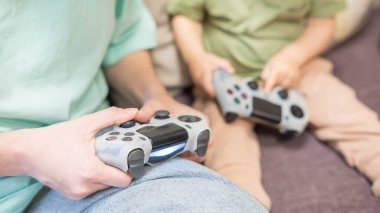 Beyaz kadın küçük oğluna konsol oynamayı öğretiyor. Oyun kolu ellerinde.