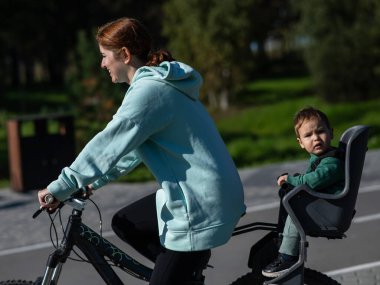 Küçük oğluyla bisiklete binen beyaz bir kadın arkasında çocuk koltuğunda oturuyor.
