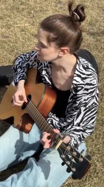 Genç beyaz kadın çimenlerde otururken gitar çalıp şarkı söylüyor.