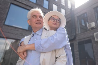 Şapkalı ve gözlüklü bir kadın beyaz ceketli yaşlı bir adama arkadan sarılıyor. Olgun insanların romantik ilişkileri