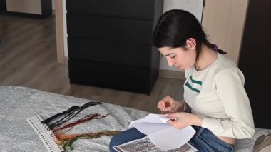 Genç beyaz kadın yatakta otururken çapraz dikiş atıyor.