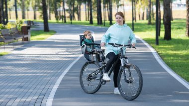 Küçük oğluyla bisiklete binen beyaz bir kadın arkasında çocuk koltuğunda oturuyor.
