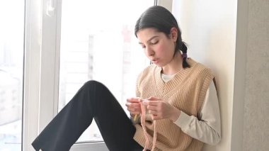 Genç beyaz kadın pencere eşiğinde otururken örgü örüyor.