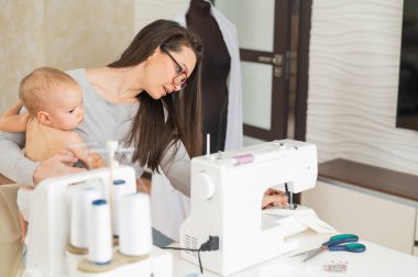 Genç bir kadın mutfakta dikiş dikiyor ve küçük bir çocuğu tutuyor. Annem küçük oğluna dikiş makinesi dikmeyi öğretiyor. Kendi kendini izole etme ödevi.