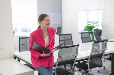 Pembe ceketli gülümseyen iş kadını ofiste evrak okuyor.