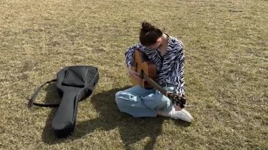 Genç beyaz kadın çimenlerde otururken gitarını akort ediyor.