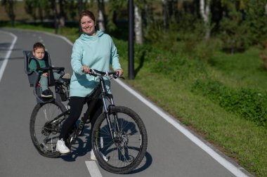 Küçük oğluyla bisiklete binen beyaz bir kadın arkasında çocuk koltuğunda oturuyor.