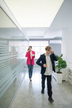 İki meslektaş ofiste yürüyor ve telefonda konuşuyorlar.