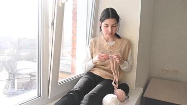 Genç beyaz kadın pencere eşiğinde otururken örgü örüyor.