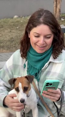 Beyaz kadın Jack Russell Terrier 'ını gezdirirken akıllı telefon kullanıyor.