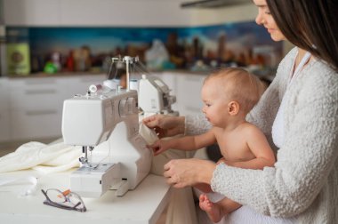 Genç bir kadın mutfakta dikiş dikiyor ve küçük bir çocuğu tutuyor. Annem küçük oğluna dikiş makinesi dikmeyi öğretiyor. Kendi kendini izole etme ödevi.