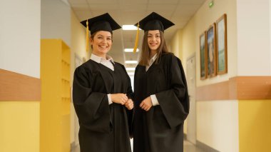 Mezuniyet elbisesi giymiş iki genç kadın üniversite koridorunda duruyor.
