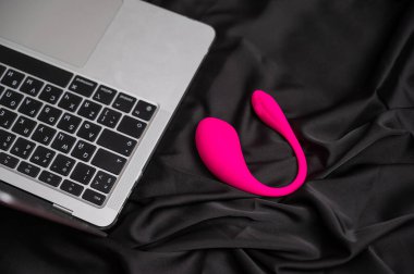 Siyah ipek çarşafta pembe kıvrımlı vibratör ve laptop.