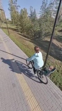 Beyaz bir kadın parkta yeni yürümeye başlayan oğluyla bisiklet sürüyor. Dikey video