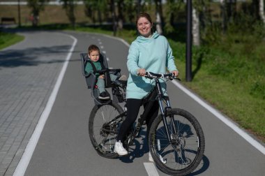 Küçük oğluyla bisiklete binen beyaz bir kadın arkasında çocuk koltuğunda oturuyor.