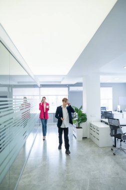 İki meslektaş ofiste yürüyor ve telefonda konuşuyorlar.