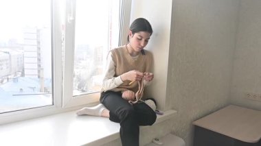 Genç beyaz kadın pencere eşiğinde otururken örgü örüyor.