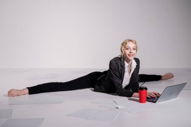 İş elbisesi giymiş bir balerin orta sicimle oturur, dizüstü bilgisayarda çalışır ve kahve içer. Bilgisayar başında esnek bir kadın çalışıyor..