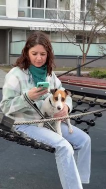 Beyaz kadın Jack Russell Terrier 'ını gezdirirken akıllı telefon kullanıyor.