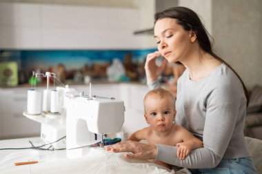 Genç bir kadın mutfakta dikiş dikiyor ve küçük bir çocuğu tutuyor. Annem küçük oğluna dikiş makinesi dikmeyi öğretiyor. Kendi kendini izole etme ödevi.