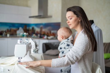 Genç bir kadın mutfakta dikiş dikiyor ve küçük bir çocuğu tutuyor. Annem küçük oğluna dikiş makinesi dikmeyi öğretiyor. Kendi kendini izole etme ödevi.