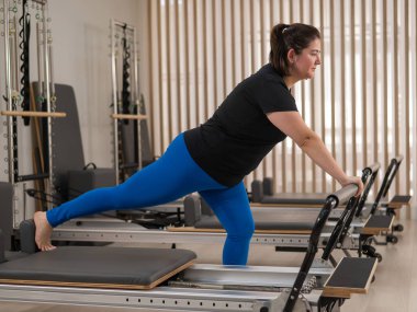 Aşırı kilolu beyaz kadın bir reformcu üzerinde pilates egzersizi yapıyor.