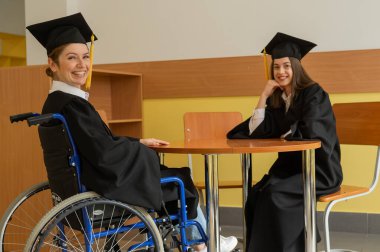 Mezuniyet elbisesi giymiş, elinde diplomasıyla tekerlekli sandalyedeki mutlu genç kadın ve kadın. Özel eğitim