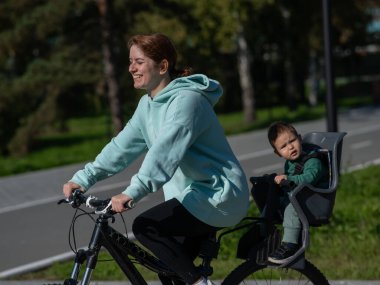 Küçük oğluyla bisiklete binen beyaz bir kadın arkasında çocuk koltuğunda oturuyor.