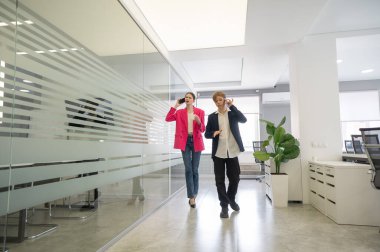 İki meslektaş ofiste yürüyor ve telefonda konuşuyorlar.