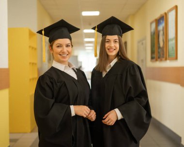 Mezuniyet elbisesi giymiş iki genç kadın üniversite koridorunda duruyor.
