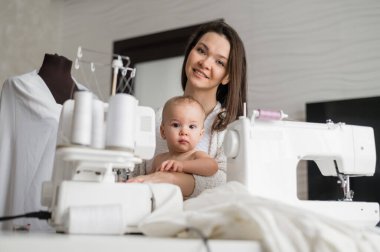 Genç bir kadın mutfakta dikiş dikiyor ve küçük bir çocuğu tutuyor. Annem küçük oğluna dikiş makinesi dikmeyi öğretiyor. Kendi kendini izole etme ödevi.