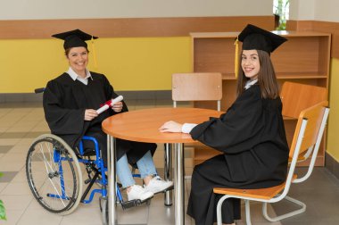 Mezuniyet elbisesi giymiş, elinde diplomasıyla tekerlekli sandalyedeki mutlu genç kadın ve kadın. Özel eğitim