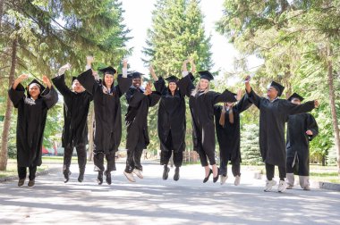 Mezuniyet elbiseleriyle dışarıda neşe içinde zıplayan çok uluslu gençler.