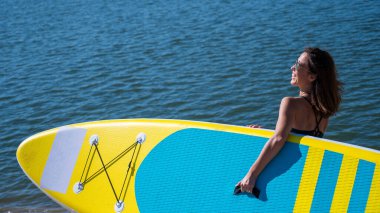 Beyaz bir kadın sahilde yürüyor ve şehirdeki nehirde bir yiyecek panosu taşıyor. Yaz sporu