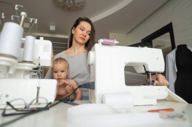 Genç bir kadın mutfakta dikiş dikiyor ve küçük bir çocuğu tutuyor. Annem küçük oğluna dikiş makinesi dikmeyi öğretiyor. Kendi kendini izole etme ödevi.