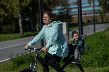 Beyaz kadın bisiklete biniyor. Yanında da oğlu var. Parkta çocuk koltuğunda oturuyor.