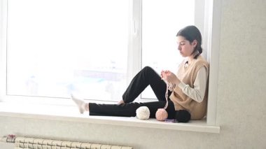 Genç beyaz kadın pencere eşiğinde otururken örgü örüyor.