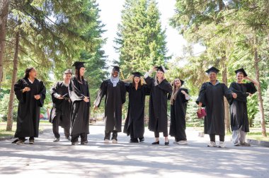 Mezuniyet elbiseleriyle dışarıda neşe içinde zıplayan çok uluslu gençler.