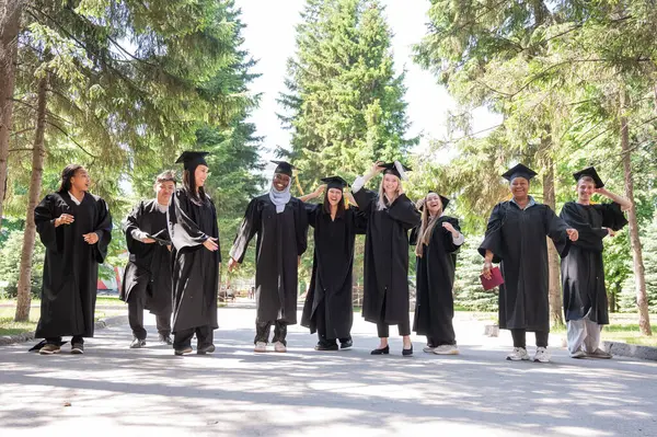 Mezuniyet elbiseleriyle dışarıda neşe içinde zıplayan çok uluslu gençler.