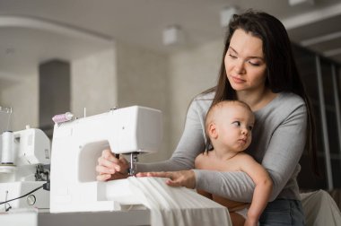 Genç bir kadın mutfakta dikiş dikiyor ve küçük bir çocuğu tutuyor. Annem küçük oğluna dikiş makinesi dikmeyi öğretiyor. Kendi kendini izole etme ödevi.