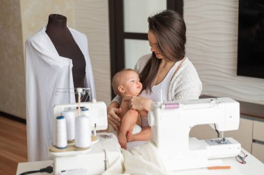 Genç bir kadın mutfakta dikiş dikiyor ve küçük bir çocuğu tutuyor. Annem küçük oğluna dikiş makinesi dikmeyi öğretiyor. Kendi kendini izole etme ödevi.