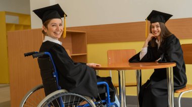 Mezuniyet elbisesi giymiş, elinde diplomasıyla tekerlekli sandalyedeki mutlu genç kadın ve kadın. Özel eğitim