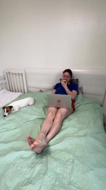 Beyaz kadın yatağında uzanıp dizüstü bilgisayarda film izliyor ve elma yiyor.