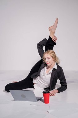 İş elbisesi giymiş çıplak ayaklı balerin dizüstü bilgisayara poz veriyor ve kahve içiyor. Bilgisayar başında esnek bir kadın çalışıyor..