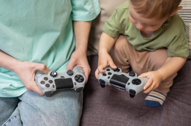 Beyaz kadın küçük oğluna konsol oynamayı öğretiyor. Oyun kolu ellerinde.