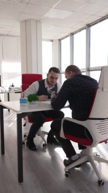 Ofiste bir masada oturan ve akıllı bir telefona bakan iki beyaz adam.