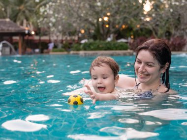 Beyaz bir kadın tatilde havuzda yüzerken oğlunu kucağına alıyor. Annesi küçük çocuğa yüzmeyi öğretiyor.