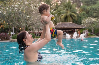 Beyaz bir kadın tatilde havuzda yüzerken oğlunu kucağına alıyor. Annesi küçük çocuğa yüzmeyi öğretiyor.